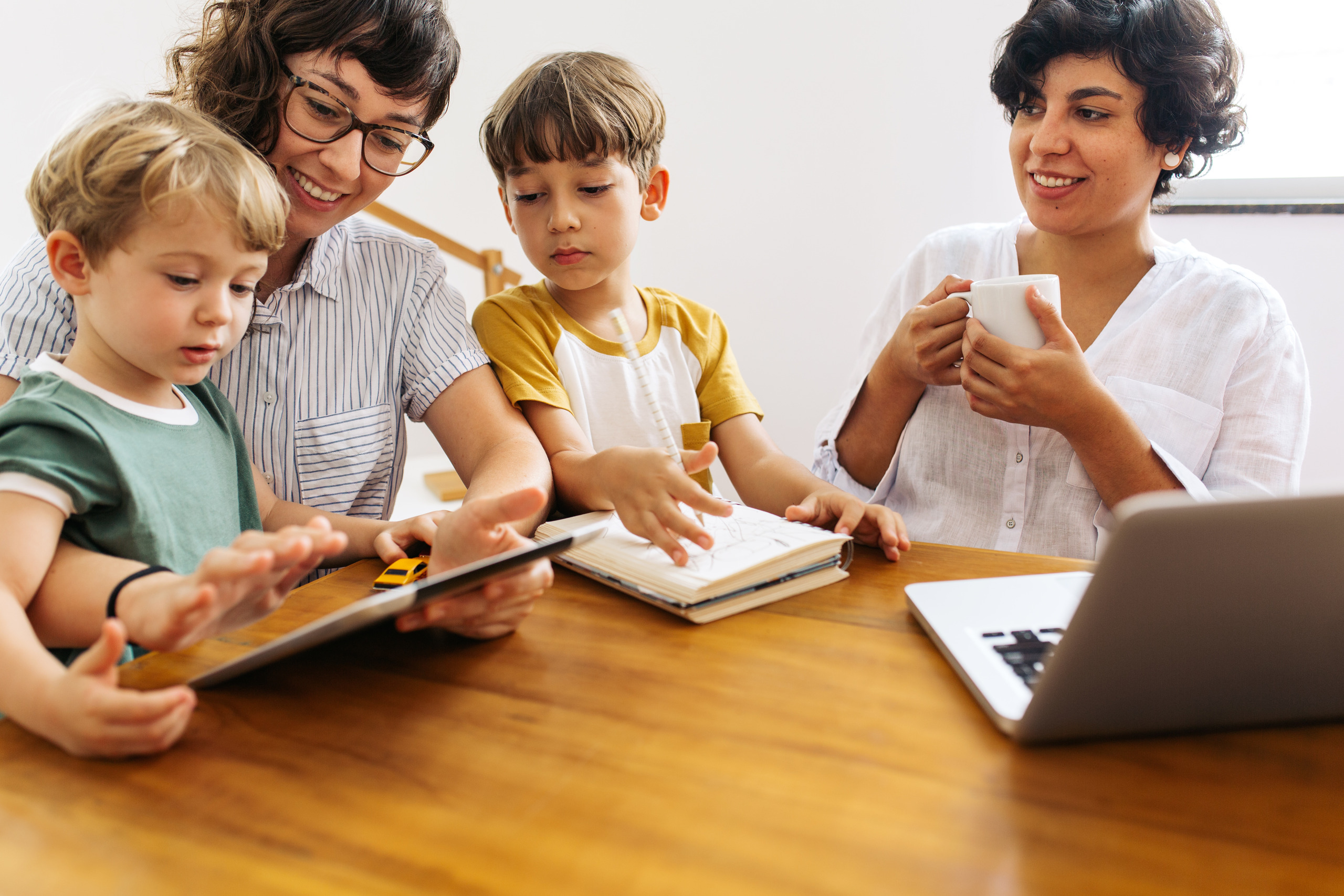 A family sits together at a wooden table. One mom holds a coffee cup and smiles at the other, who is holding a tablet for one child as the other looks over her shoulder and draws in a notebook.