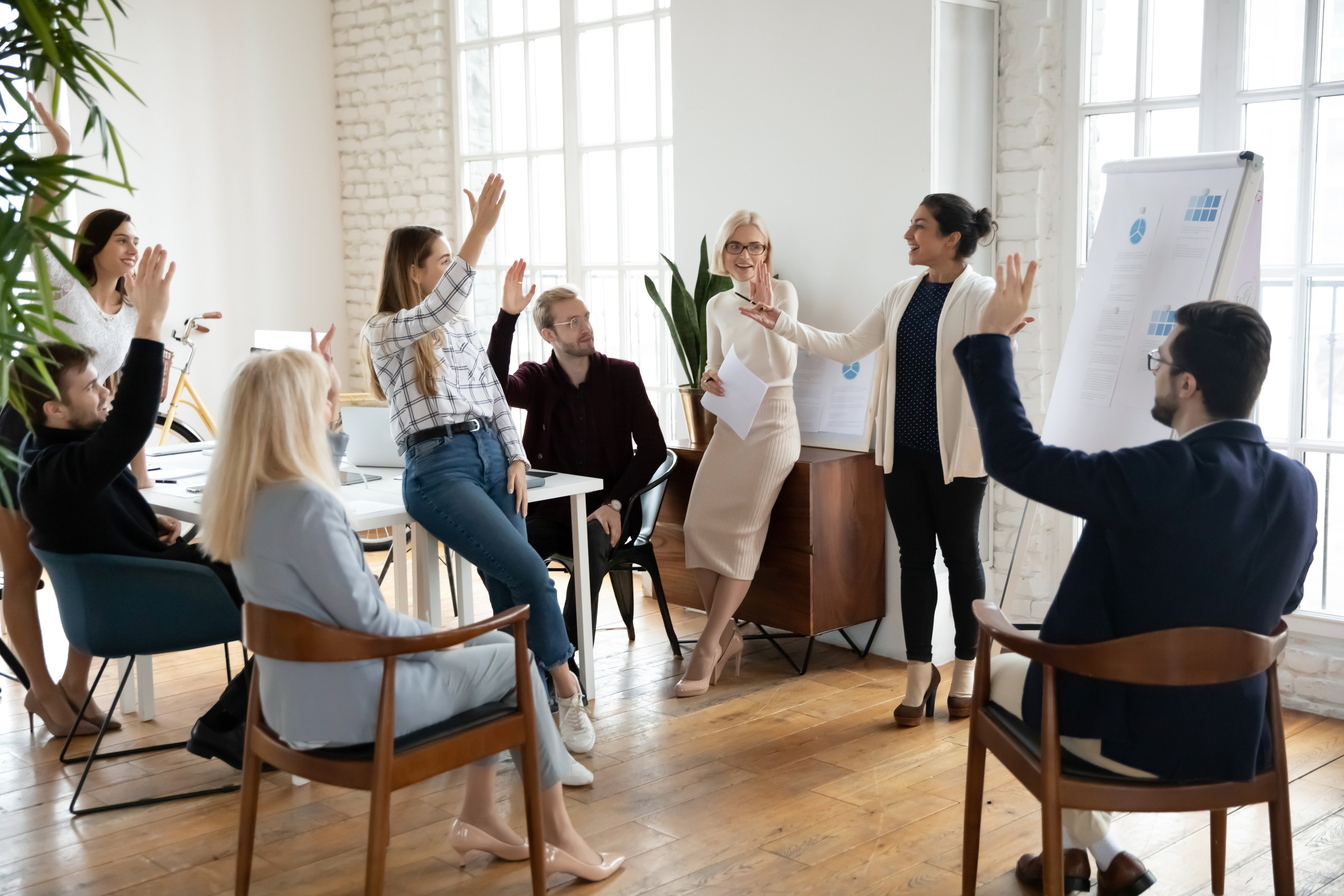 A group of people in business clothes sit and stand together in a meeting as one of them presents charts on an easel. All of them are raising their hands as if asking questions or voting on a question